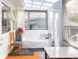 A sunroom with a white covered sofa and pillows a wooden chair flowers on a small table and a glass door at Haere Mai Reefton Heritage Holiday Home in Reefton