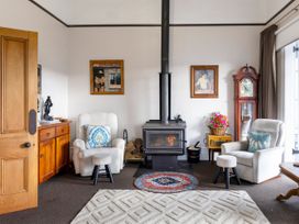 A sitting area with two armchairs a wood-burning stove a wooden cabinet a grandfather clock and framed paintings at Haere Mai Reefton Heritage Holiday Home in Reefton
