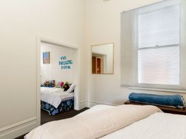 A bedroom with a bed blanket rolled on a wooden bench under a window and a view into a smaller room with a bed and plush toys at Haere Mai Reefton Heritage Holiday Home in Reefton