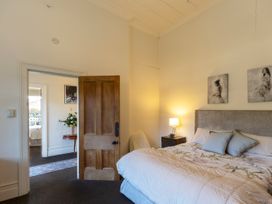 A bedroom with a bed and two pillows a nightstand with a lamp an open wooden door showing another room with a table and flowers at Haere Mai Reefton Heritage Holiday Home in Reefton