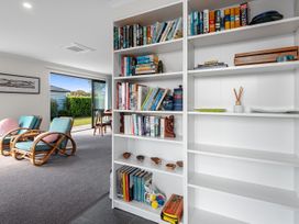 A living room with a white bookshelf filled with books and decorative items beside two blue cushioned armchairs with pink pillows at Bennett's Bach - Waipu Holiday Home in Waipu