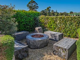 An outdoor seating area with wooden benches around a stone fire pit surrounded by green hedges at Bennetts Bach Waipu Holiday Home in Waipu