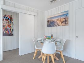 A dining area with a glass table and four white chairs with wooden legs a colorful portrait on the wall and a beach landscape picture at Boulevard Bliss - Pauanui Holiday Home in Pauanui