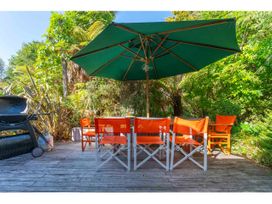 An outdoor wooden deck with a green umbrella shading orange chairs and a table surrounded by greenery and a barbecue grill Peaceful Outlook - Hatepe Holiday Home Hatepe