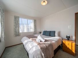 A bedroom with a bed covered in white and pink bedding three dark blue pillows rolled towels on top a wooden nightstand with a black lamp and two windows with curtains at Cosy Crib - Te Anau Holiday Home in Te Anau