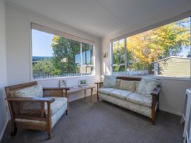 A small sitting room with a wooden armchair and sofa with floral cushions and a rectangular bench by large windows at Relaxing Kiwi Bach - Te Anau Holiday Home in Te Anau
