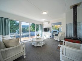 A living room with white furniture including armchairs and a coffee table a television on a stand large windows with curtains and a wood stove at Relaxing Kiwi Bach Te Anau Holiday Home in Te Anau