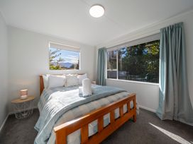 A bedroom with a wooden bed frame a side table with a lamp and large windows with curtains at Relaxing Kiwi Bach - Te Anau Holiday Home in Te Anau