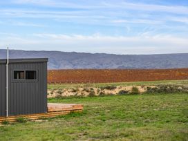 A dark gray building with a window and wooden deck next to a grassy field with vineyard and hills in the background at Alex Vineyard - Alexandra Holiday Home in Alexandra