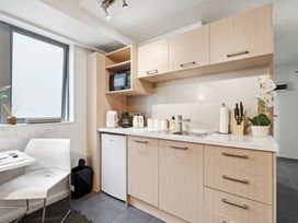 A kitchen with light wood cabinets a white countertop a sink a microwave kettle coffee maker toaster and a white chair by a window at Cityscape on White - Auckland Central Studio in Auckland Central