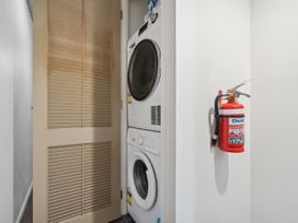 A laundry area with a stacked washer and dryer beside a beige louvered door and a red fire extinguisher mounted on a white wall at Cityscape on White Auckland Central Studio in Auckland Central
