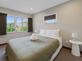 A bedroom with a bed covered with a green bedspread, two rolled towels on the bed, a side table with a lamp, and windows with curtains at Bell Cottage - Oakura Holiday Home in Oakura