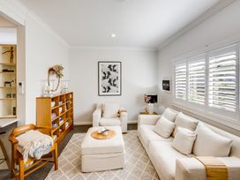 A living room with a white sofa armchair and ottoman wooden chair bookshelf and window shutters at Coastal Charm Napier Holiday Home in Napier