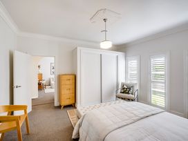 A bedroom with a bed a wooden chest of drawers a chair and three windows with shutters at Coastal Charm - Napier Holiday Home in Napier