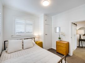 A bedroom with a bed, yellow bedside table with lamp, wooden dresser with plant, and a window with blinds at Coastal Charm - Napier Holiday Home in Napier