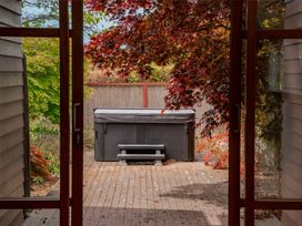 A wooden deck with a covered hot tub and small steps surrounded by trees with red and green leaves viewed through glass doors at Elderberry Wine - Wanaka Holiday Home Wanaka