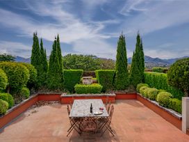 An outdoor patio area with a tiled table and metal chairs surrounded by shaped green hedges and trees with mountains in the background at Elderberry Wine - Wanaka Holiday Home in Wanaka