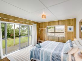 A bedroom with a bed covered in blue and white quilt next to sliding glass doors and a window with wooden panel walls at Sunset Haven - Mahia Holiday Home in Mahia Peninsula
