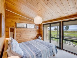 A bedroom with a bed covered by a patterned quilt and pillows a wooden ceiling and walls a hanging round paper light a chair with towels and sliding glass doors opening to a porch with outdoor views at Sunset Haven - Mahia Holiday Home in Mahia Peninsula