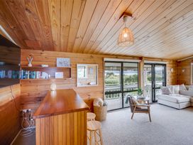 A living room with wooden walls and ceiling a wooden bar with stools a gray armchair and a white sectional sofa near sliding glass doors at Sunset Haven - Mahia Holiday Home in Mahia Peninsula