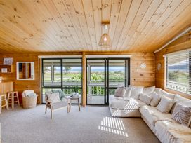 A living room with a beige sectional sofa and an armchair near sliding glass doors showing an outdoor view at Sunset Haven - Mahia Holiday Home in Mahia Peninsula