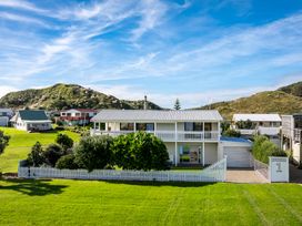 A two story house with a white picket fence and green bushes in front on a grassy lawn with hills in the background at Sunset Haven - Mahia Holiday Home in Mahia Peninsula