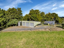 A outdoor area with a green grass field a gravel driveway a building with three closed garage doors trees in the background and a clear sky at Glenmohr Cabin - Waipu Holiday Retreat in Waipu