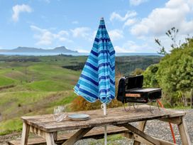 A wooden picnic table with a folded blue striped umbrella a small barbecue grill two glasses and plates outdoors with green hills and ocean in the background at Glenmohr Cabin - Waipu Holiday Retreat in Waipu