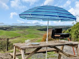 A wooden picnic table with glasses plates a barbecue and a blue striped umbrella overlooking green hills and a distant body of water at Glenmohr Cabin Waipu Holiday Retreat in Waipu