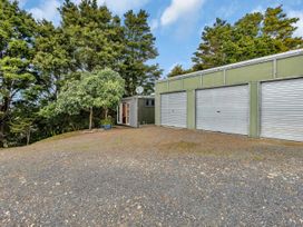 A gravel driveway with three closed garage doors and a green storage shed with open double doors surrounded by trees at Glenmohr Cabin - Waipu Holiday Retreat in Waipu