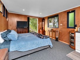 A bedroom with a bed covered in blue bedding a television on a wooden dresser and windows showing trees outside at Glenmohr Cabin - Waipu Holiday Retreat in Waipu