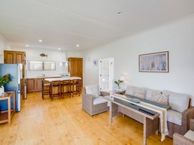 An open plan kitchen and living room with wooden cabinets stools wicker sofas and a glass table at Mahia Bliss - Surf & Serenity Holiday Home in Mahia Peninsula