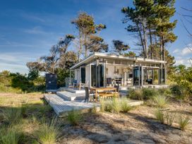 A small house with outdoor wooden deck and chairs surrounded by tall trees and grass at Mahanga Dunes Retreat - Mahia Holiday Home Mahia Peninsula