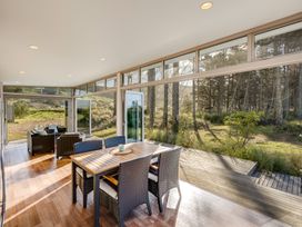 A dining area with a wooden table and wicker chairs next to a seating area with armchairs in a room with large glass windows at Mahanga Dunes Retreat - Mahia Holiday Home in Mahia Peninsula