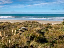 A beach view with ocean waves and grassy dunes with a round wooden picnic table at Mahanga Dunes Retreat - Mahia Holiday Home in Mahia Peninsula