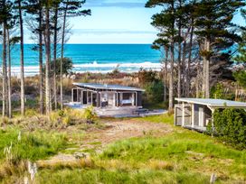A beach house with glass windows near trees and grass with ocean waves in the background at Mahanga Dunes Retreat Mahia Holiday Home Mahia Peninsula