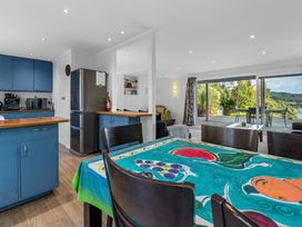 An open kitchen and dining area with blue cabinets and a table with a colorful tablecloth next to a living room with a sliding glass door leading to a deck with outdoor furniture and greenery outside at Okere Jetty Escape -  Rotoiti Holiday Home in Okere Falls