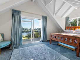 A bedroom with a wooden bed blue curtains a blue chair and sliding glass doors opening to a balcony at Okere Jetty Escape - Rotoiti Holiday Home in Okere Falls