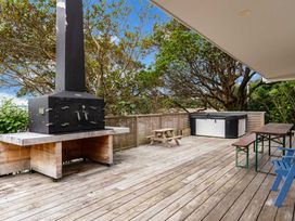 An outdoor wooden deck with a large black fireplace oven a hot tub a picnic table a long wooden table with benches and blue chairs at Heron's Hideaway - Snells Beach Holiday Home in Snells Beach