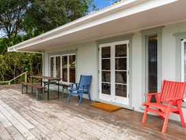 An outdoor wooden deck with a picnic table bench and two chairs in front of a house with glass doors and windows at Heron's Hideaway - Snells Beach Holiday Home in Snells Beach