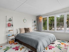 A bedroom with a bed with pillows a gray blanket two white ladder shelves a wicker lamp floral rug and large windows showing trees at Heron's Hideaway - Snells Beach Holiday Home in Snells Beach