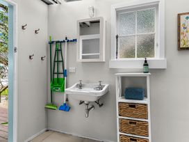 A utility room with a wall sink mop and broom hooks a window and storage unit with baskets at Heron's Hideaway - Snells Beach Holiday Home in Snells Beach