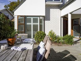 An outdoor patio area with a wooden dining table set with placemats glasses and a jug in front of a white brick house with plants and shrubs at Stylish Fendalton Townhouse Prime Location Christchurch