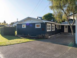 A single story house with dark siding white trim windows and a driveway with a tree in the foreground at Waterside Bliss - Taupo Holiday Home in Taupo Central