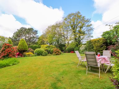 A garden with lawn chairs and a table with a pink tablecloth surrounded by trees and bushes at Trevow in Camborne