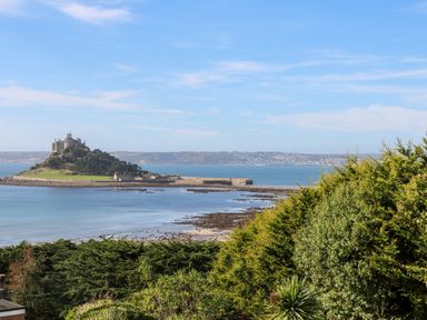 An island with a castle and trees by the sea viewed from a shoreline with bushes and greenery at Trevow in Camborne