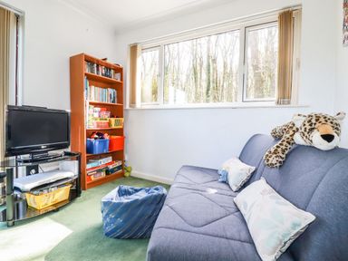 A small room with a blue sofa with pillows and a stuffed leopard toy a wooden bookshelf with books and games a TV on a glass stand and a blue bean bag on a green carpet at Trevow in Camborne