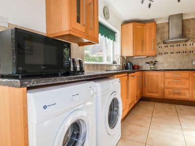 A kitchen with wooden cabinets tiled floor two white washing machines microwave toaster and window with green blind at Trevow in Camborne