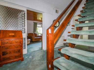 A hallway with green carpeted stairs and a wooden chest of drawers next to a doorway leading to a living room with brown leather sofas at Trevow in Camborne