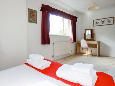A bedroom with a bed with folded towels a window with red curtains a dressing table with a chair and framed art on the walls at Trevow in Camborne